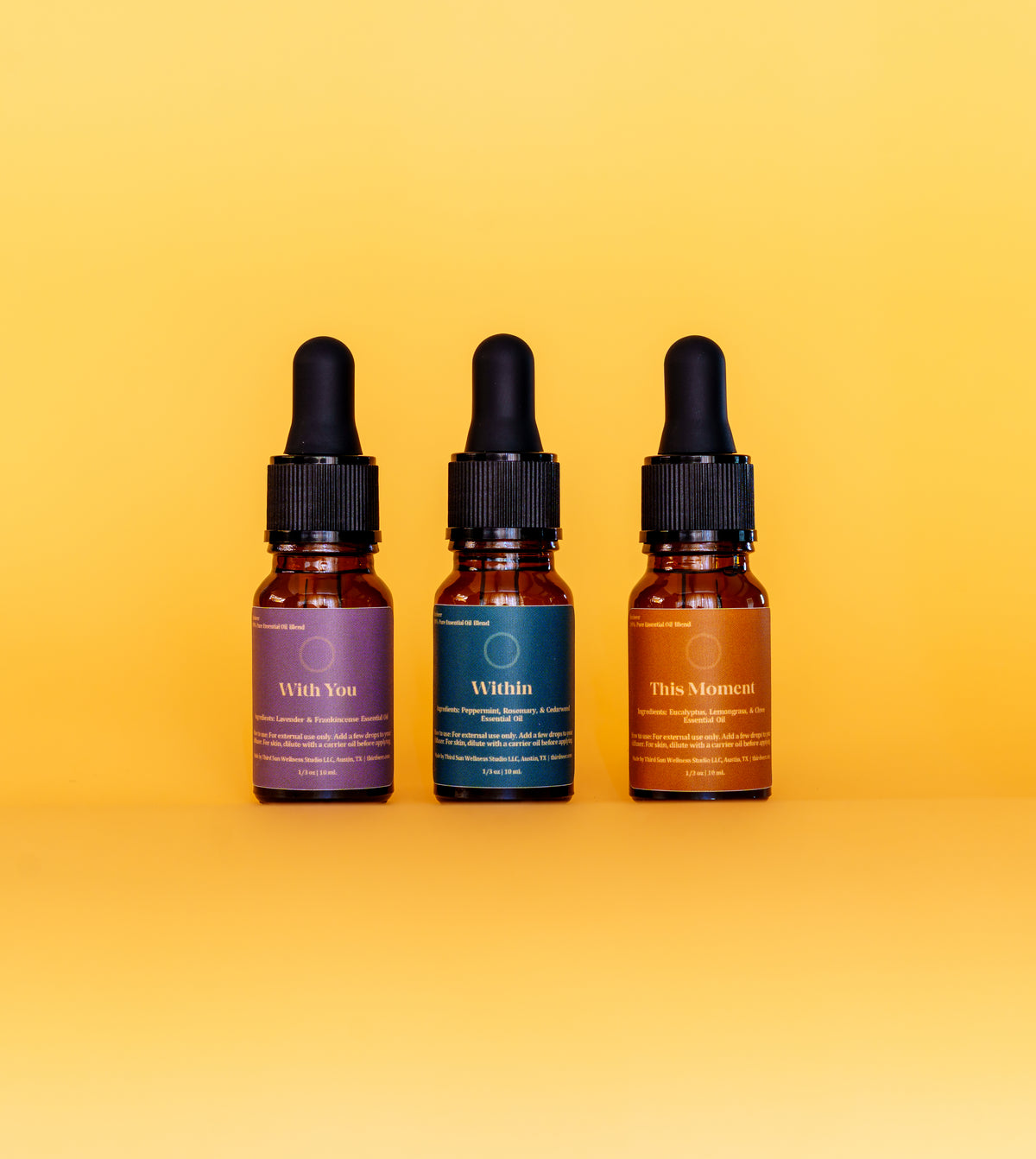 Three amber glass dropper bottles with black caps are lined up against a solid yellow background. The bottles have purple, teal, and orange labels with white text, labeled 'With You', 'Within', and 'This Moment' respectively.