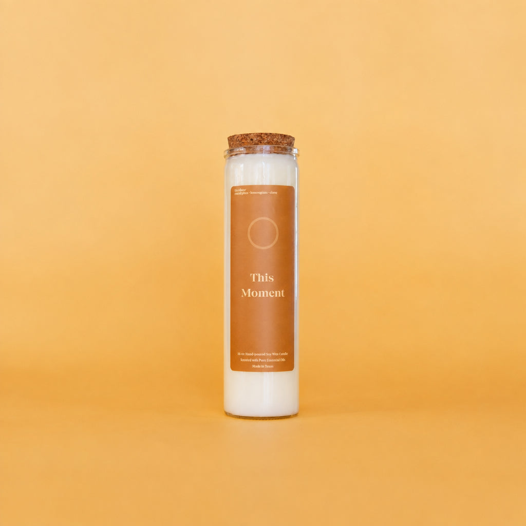 A tall glass jar candle with white wax and a cork stopper, featuring a brown label that reads 'This Moment' on a warm yellow background.