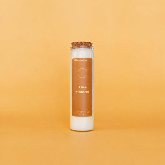 A tall glass jar candle with white wax and a cork stopper, featuring a brown label that reads 'This Moment' on a warm yellow background.