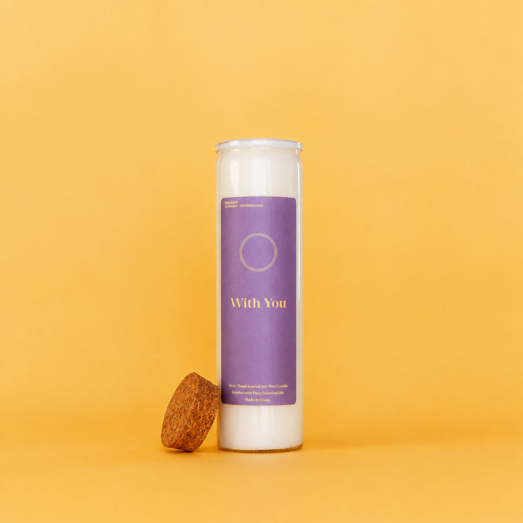 A tall white glass jar candle with a purple label reading 'With You' stands on a warm yellow-orange background, with a cork stopper resting beside the base.