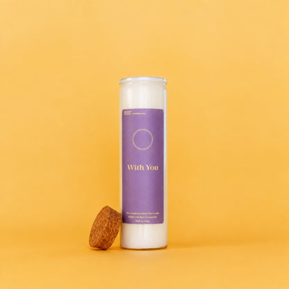 A tall white glass jar candle with a purple label reading 'With You' stands on a warm yellow-orange background, with a cork stopper resting beside the base.