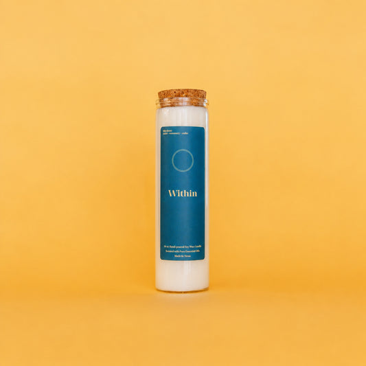 A tall white soy wax candle in a clear glass jar with a cork stopper, featuring a dark teal label that reads 'Within' on a solid yellow background.