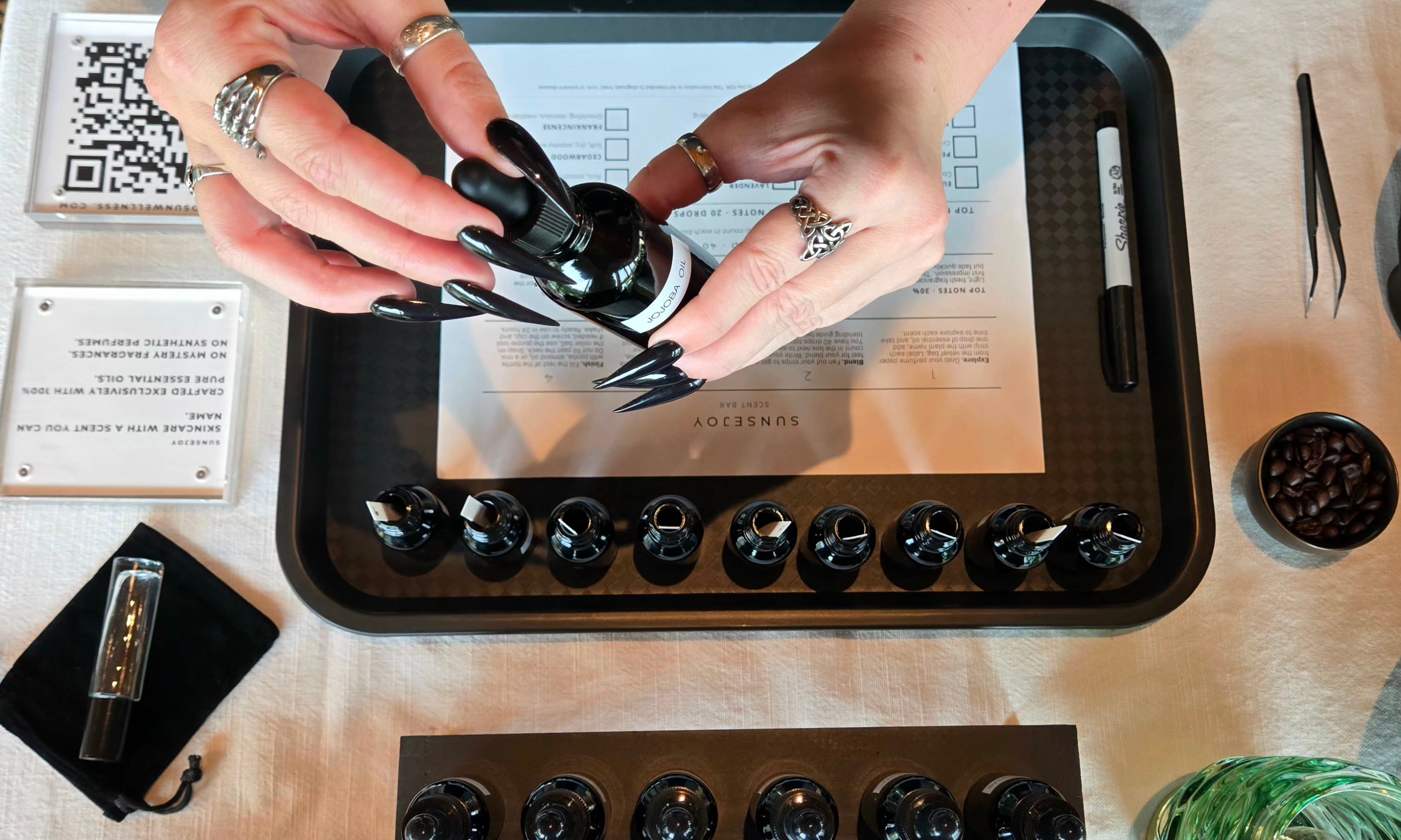 A top-down view of a person's hands holding a small black bottle with a dropper over a tray of small sample vials, likely during a fragrance or oil tasting. The scene includes a tasting form, tweezers, a pen, and other tasting accessories on a light-colored surface.